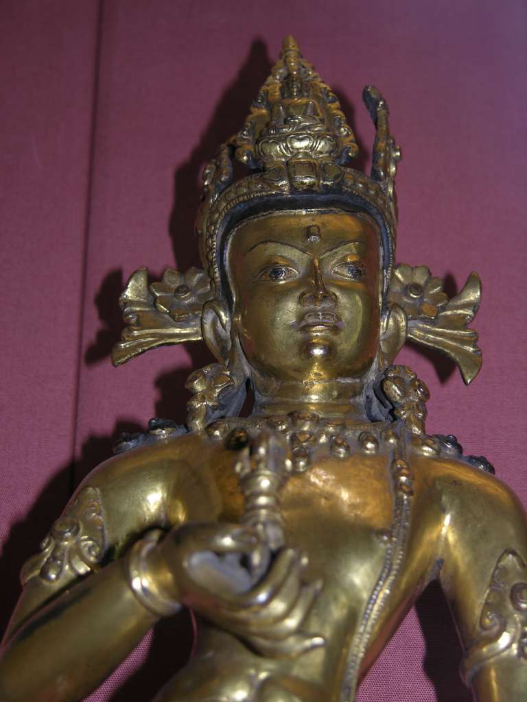 British Museum Top 20 Buddhism 16-2 Vajrasattva Close Up 16. Vajrasattva  Nepal, 15C AD, 52cm high. Here is a close up of Vajrasattva holding the vajra and bell in his hands, has elaborate jewelry, and is inset with semi-precious stones. In Vajrasattva's crown is the seated figure of the Akshobhya, the Tathagata Meditation Buddha of the East.